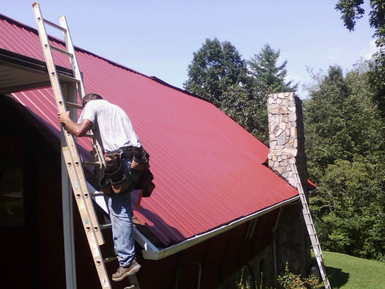 Roofer on a ladder beside a red metal roof and stone chimney — Soffit & Fascia Repair by Roofing Brothers in Maynardville